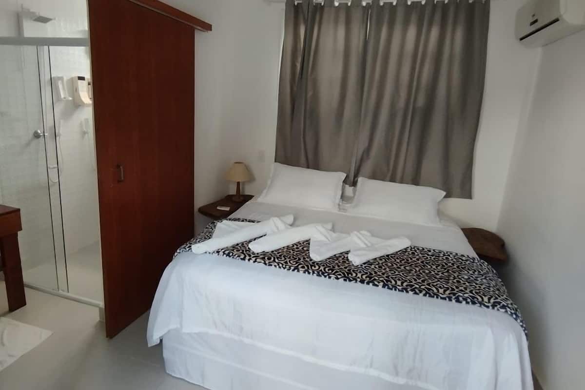 A hotel room with a bed, a nightstand with a lamp, a wooden wardrobe, and a bathroom with a glass door.