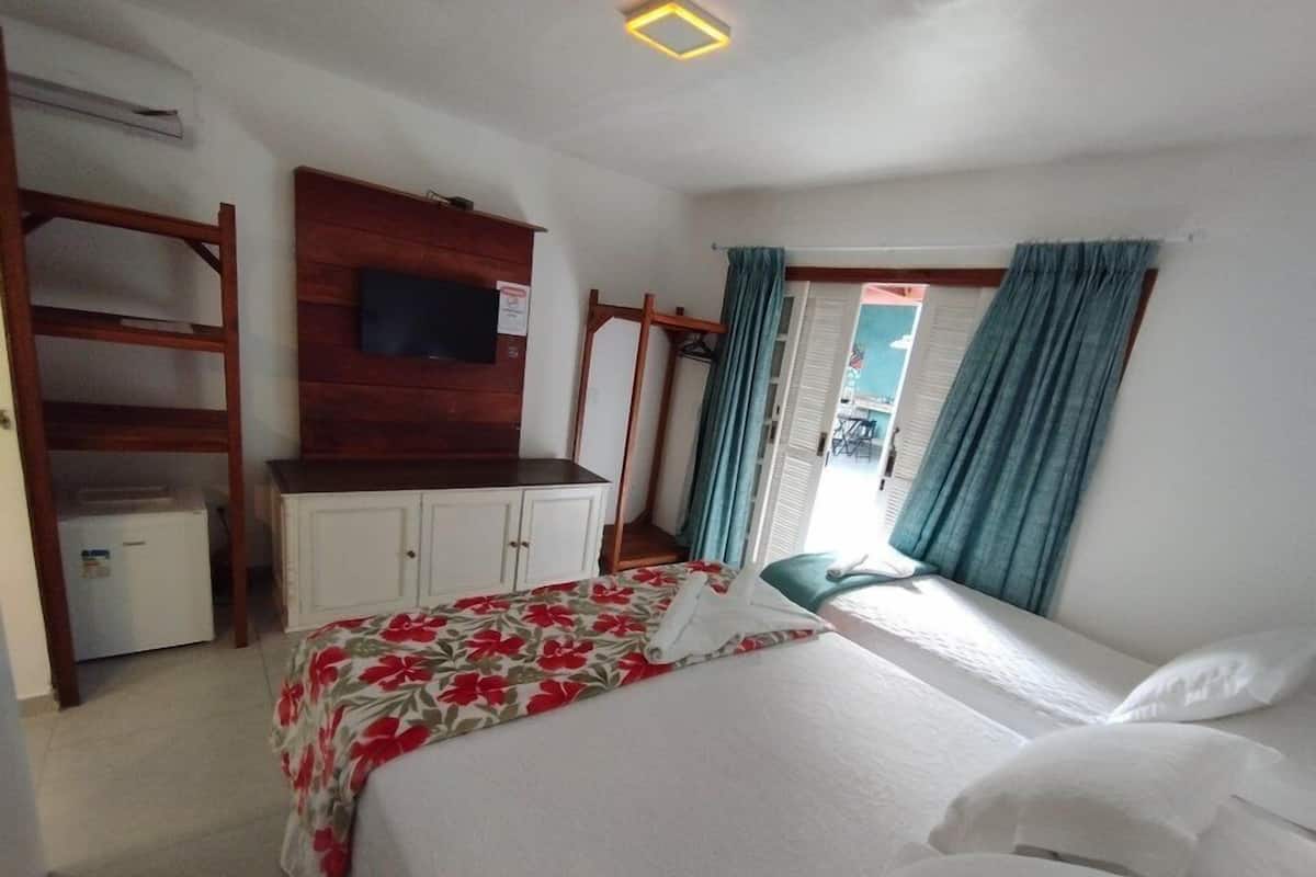 A bedroom with a bed, a TV mounted on a wooden panel, a small white cabinet, and a window with curtains.