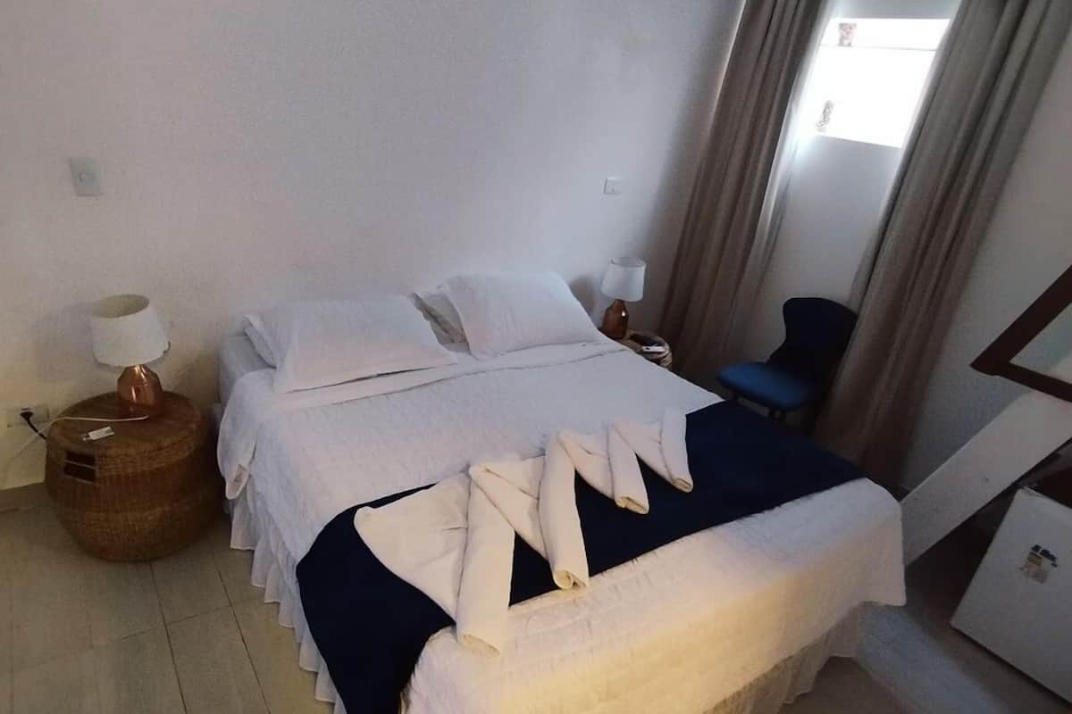 A neatly made bed with white linens and a dark blue blanket, a small round table with a lamp, a chair, and a window with curtains.