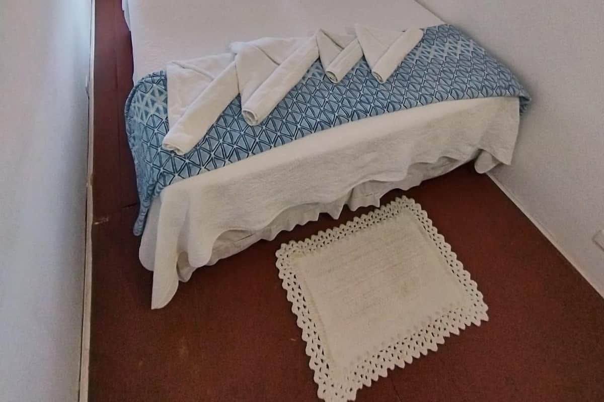 A single bed with a patterned bedspread and white pillows, a flat-screen TV mounted on the wall, and a small bedside table with a lamp.