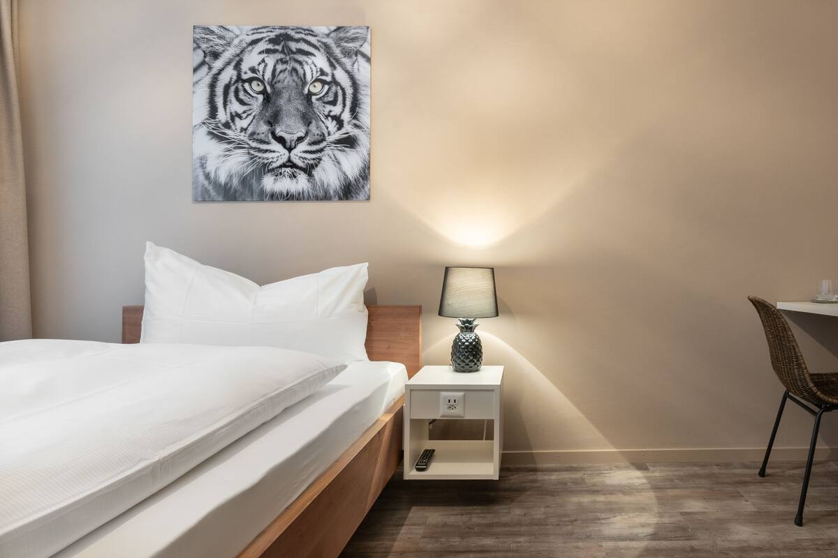 A hotel room with a bed, a nightstand with a lamp, a chair, and a wall-mounted artwork of a tiger.