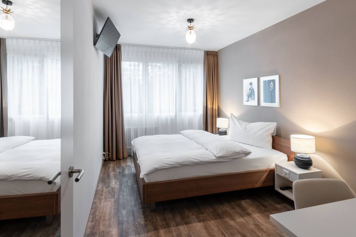 A hotel room with a wooden bed, a bedside table with a lamp, a TV mounted on the wall, and a window with sheer curtains.