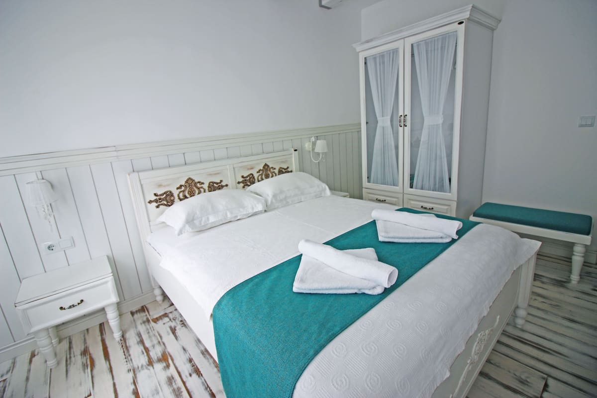 A neatly made bed with a teal bedspread and white linens, a white bedside table with drawers, a white wardrobe with a mirror, and a bench at the foot of the bed.