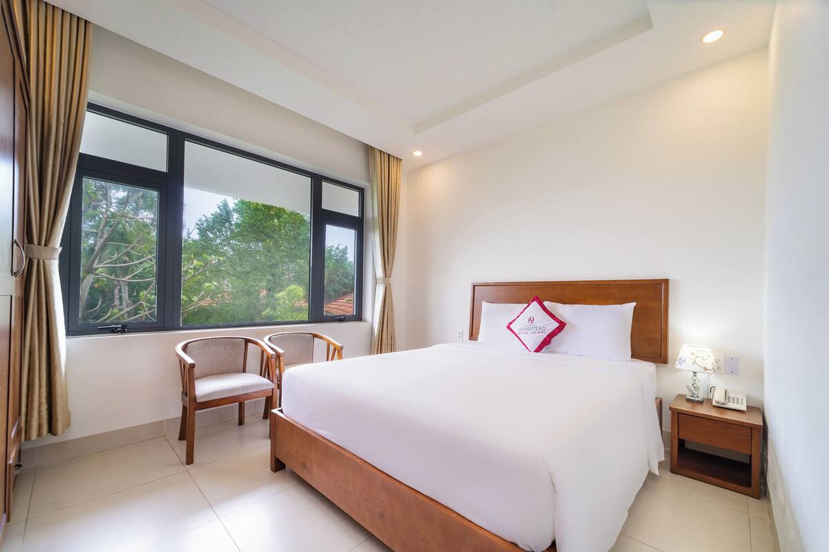 A hotel room with a large bed, a wooden nightstand with a lamp, a chair, and a window with curtains.