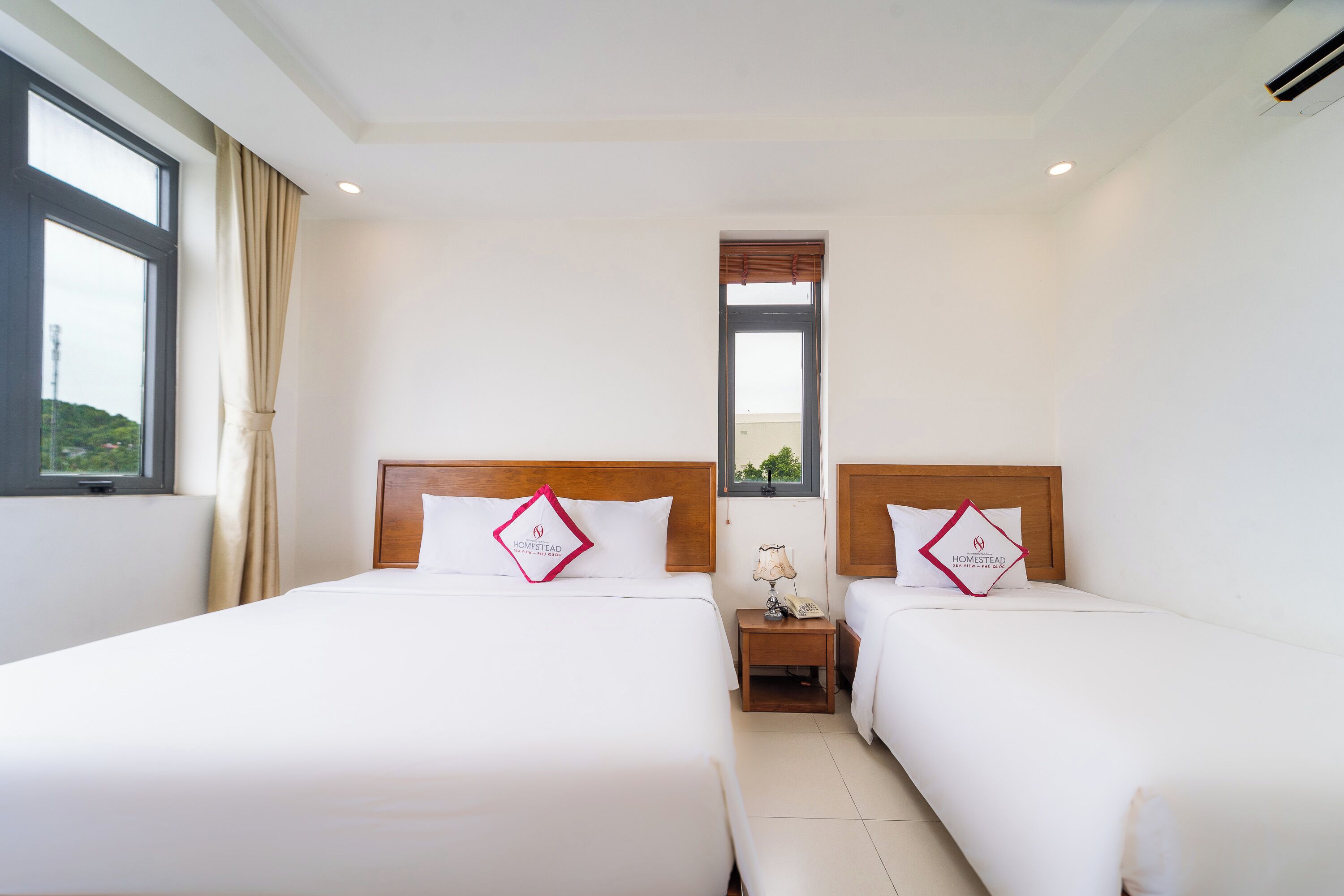 A hotel room with two beds, a window, a nightstand with a lamp, and a view of greenery outside.