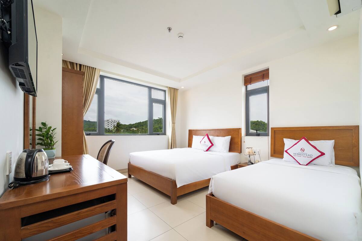A hotel room with two beds, a wooden desk, a television, and large windows.