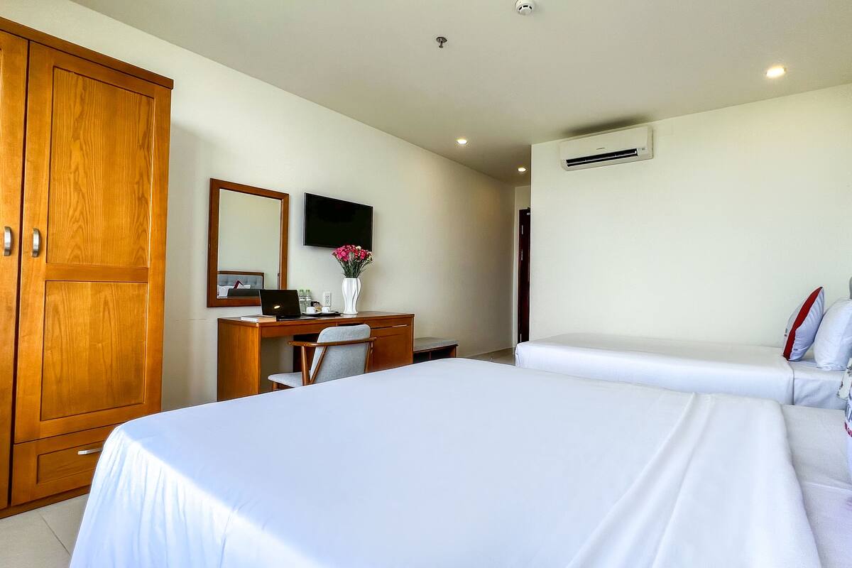 A hotel room with a large bed, a desk with a computer, a chair, a TV, and an air conditioning unit.