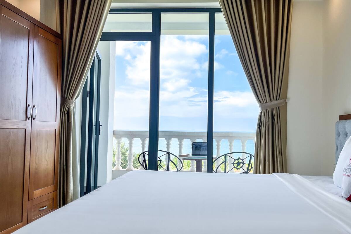A hotel room with a large bed, a view of the sea, and a balcony with a table and chairs.