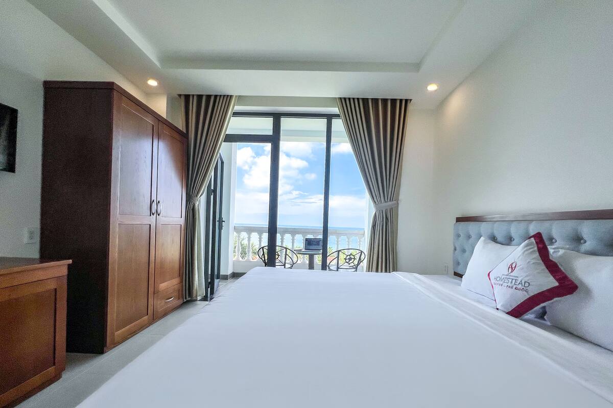 A hotel room with a large bed, a wooden wardrobe, a view of the balcony, and a table with chairs.
