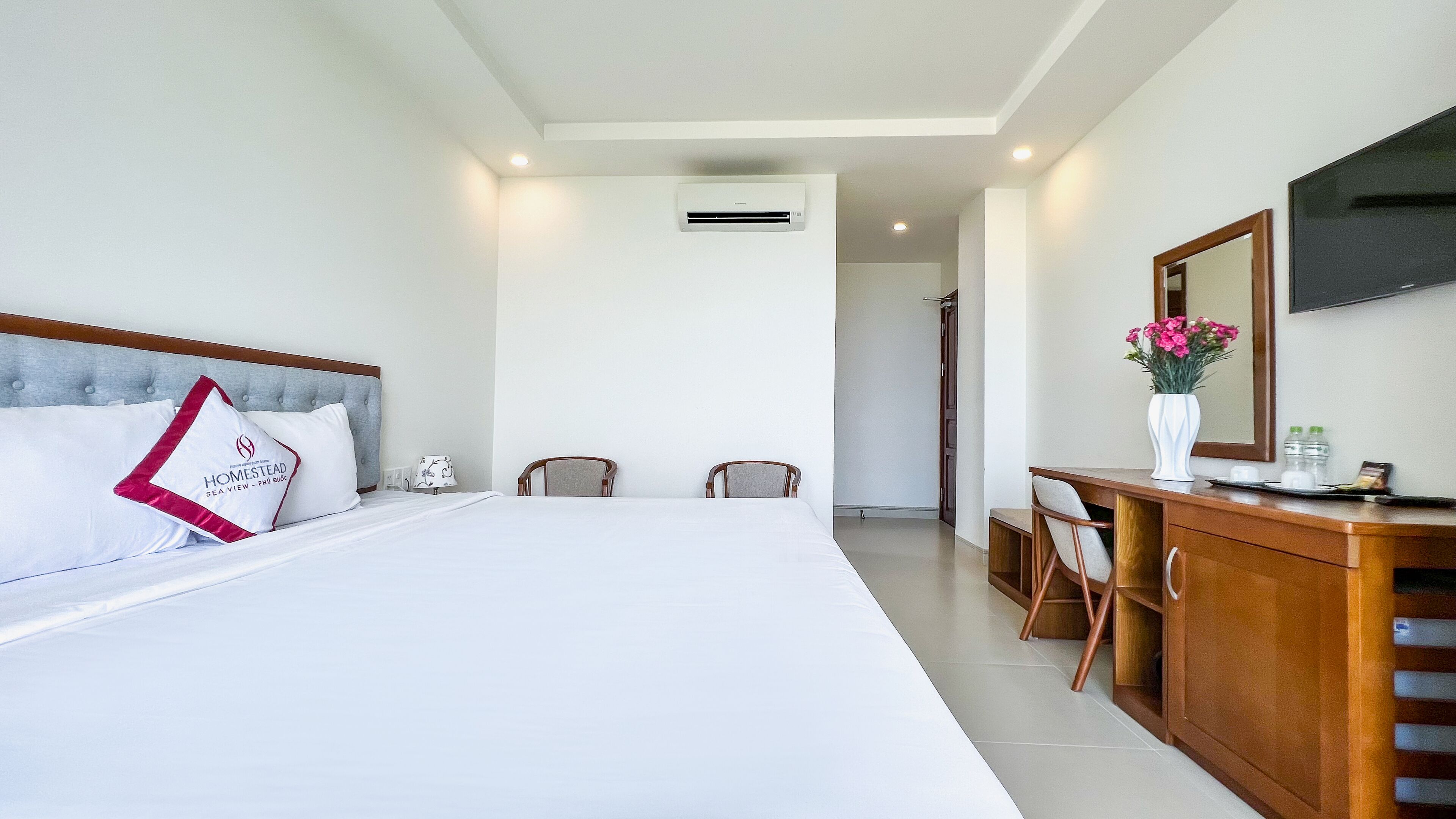A hotel room with a large bed, two chairs, a desk with a mirror, and a flat-screen TV on the wall.