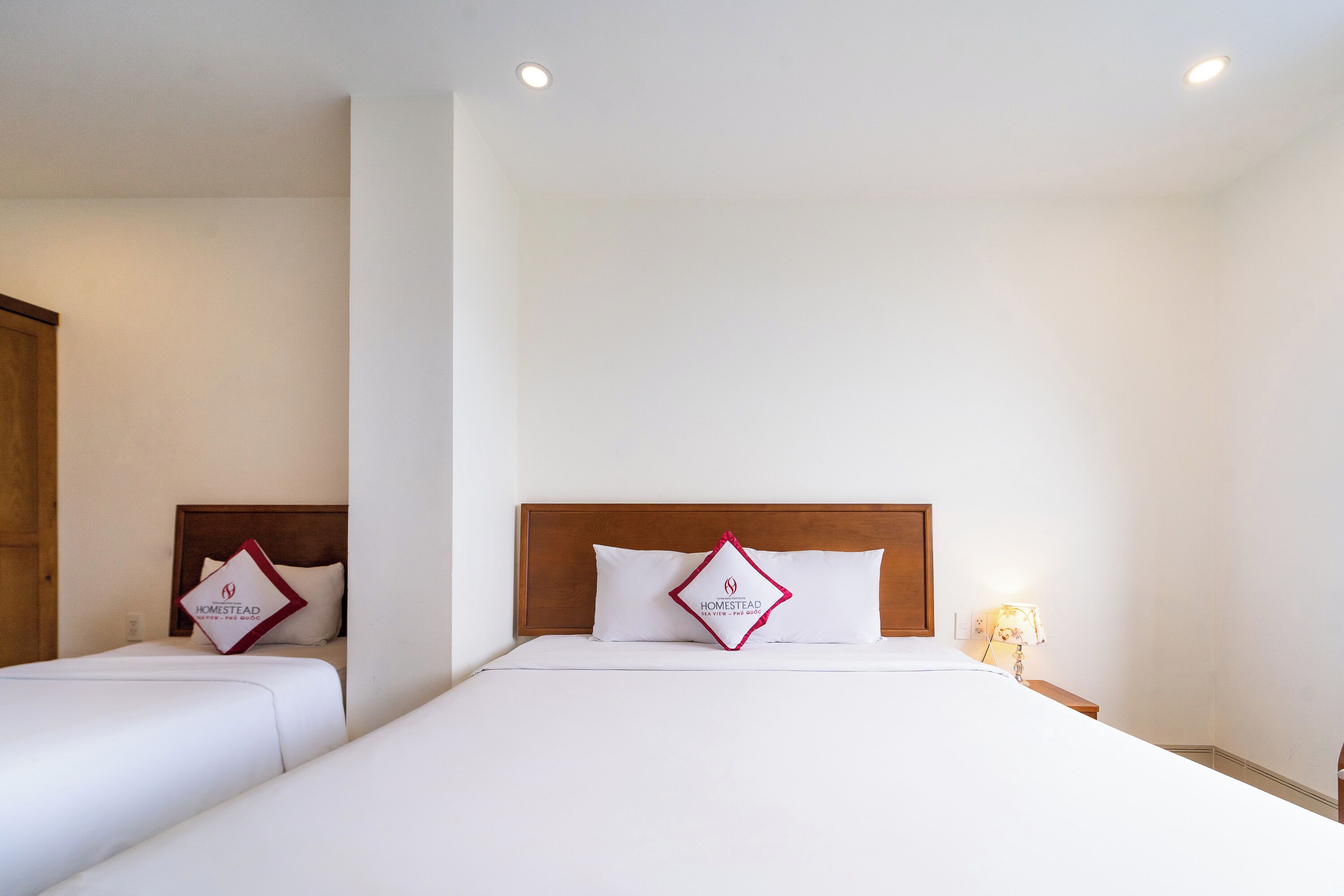 Een hotelkamer met twee bedden, elk met een houten hoofdeinde en een witte spreidek. Daartussen staat een nachtkastje met een lamp.