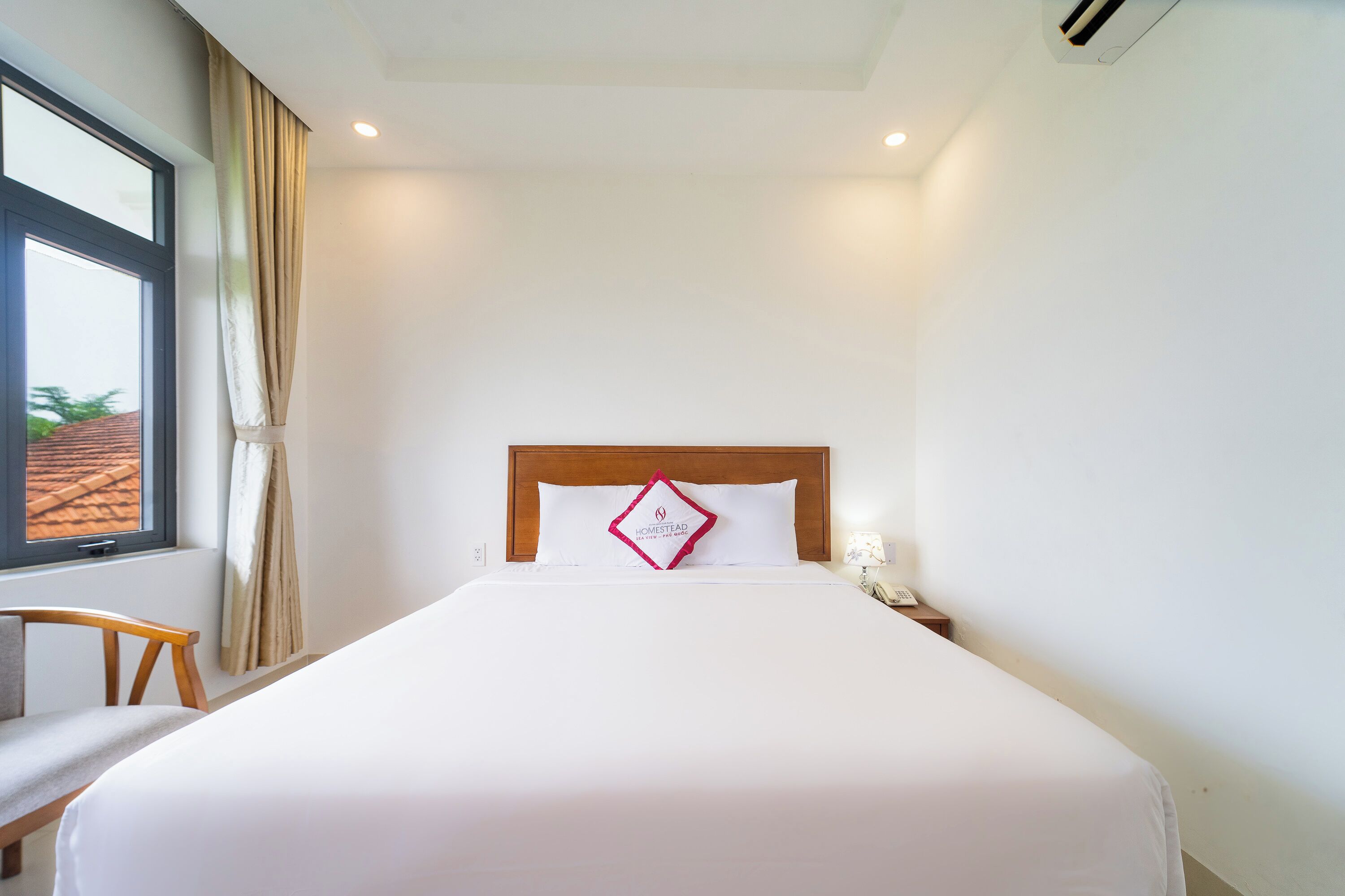 Une chambre d’hôtel avec un lit simple, une tête de lit en bois, une table de chevet avec une lampe, une chaise et une fenêtre avec des rideaux.