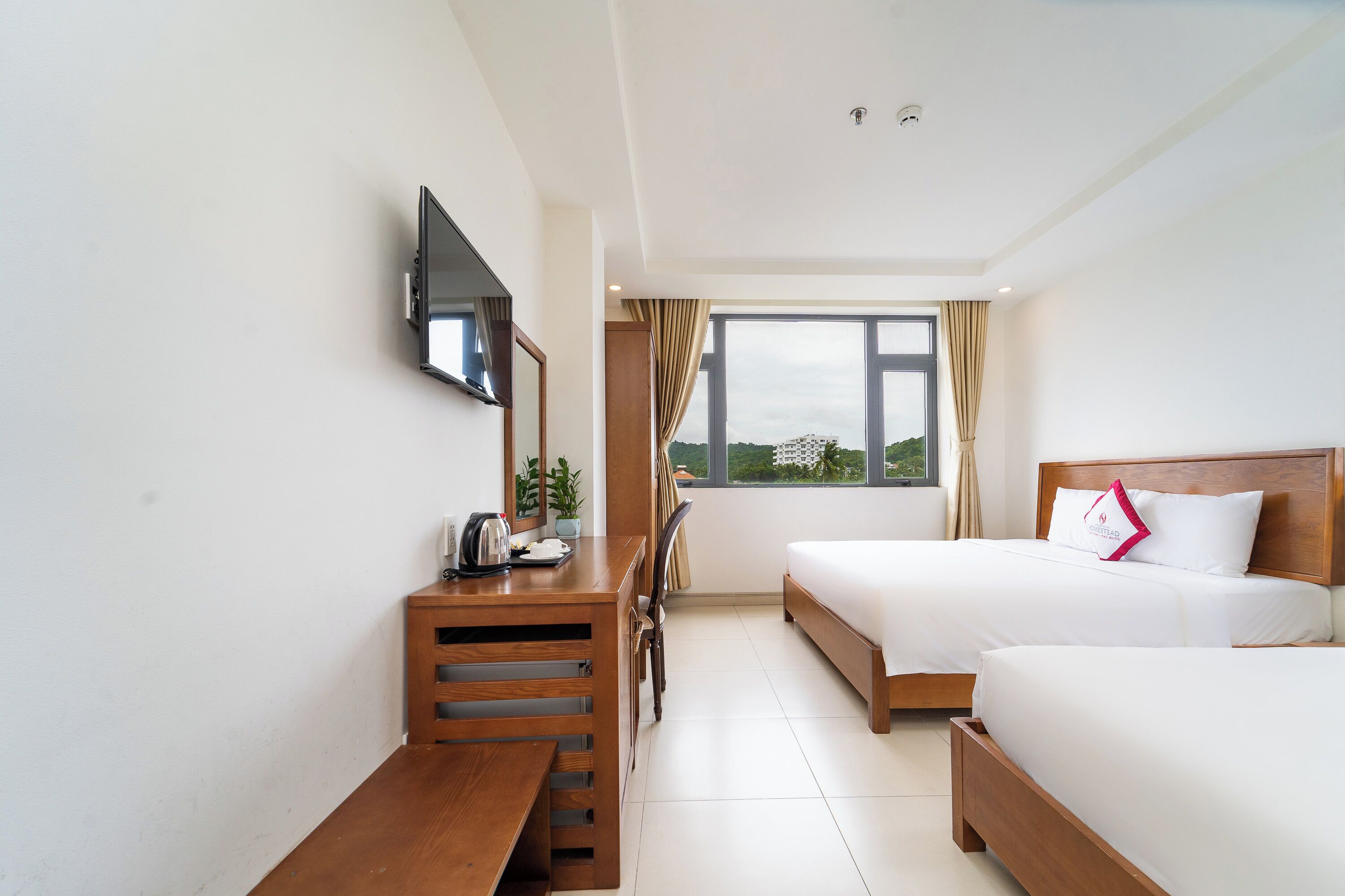 Quarto de hotel com cama, escrivaninha com cadeira, televisão na parede e janela com cortinas.
