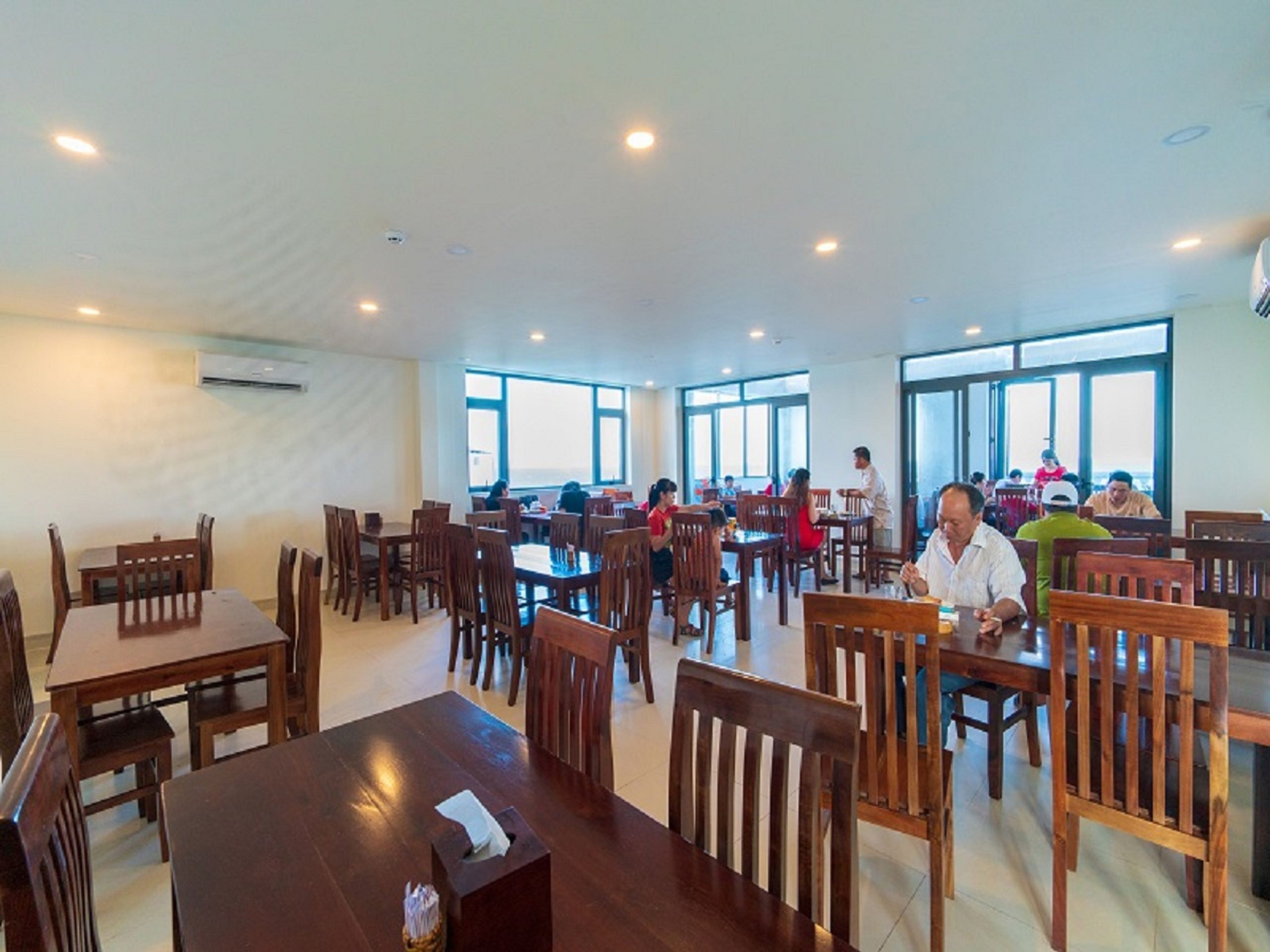 Un vaste espace dédié à la salle à manger, avec de nombreuses tables et chaises en bois, de grandes fenêtres laissant entrer la lumière naturelle et un appareil de climatisation fixé au mur.