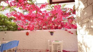 Un patio al aire libre con una mesa y sillas blancas, rodeado por un muro blanco y plantas florecientes de color rosa.