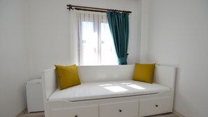 Una habitación pequeña con cama individual, paredes blancas, dos almohadas amarillas, una ventana con cortinas color turquesa y un radiador blanco.