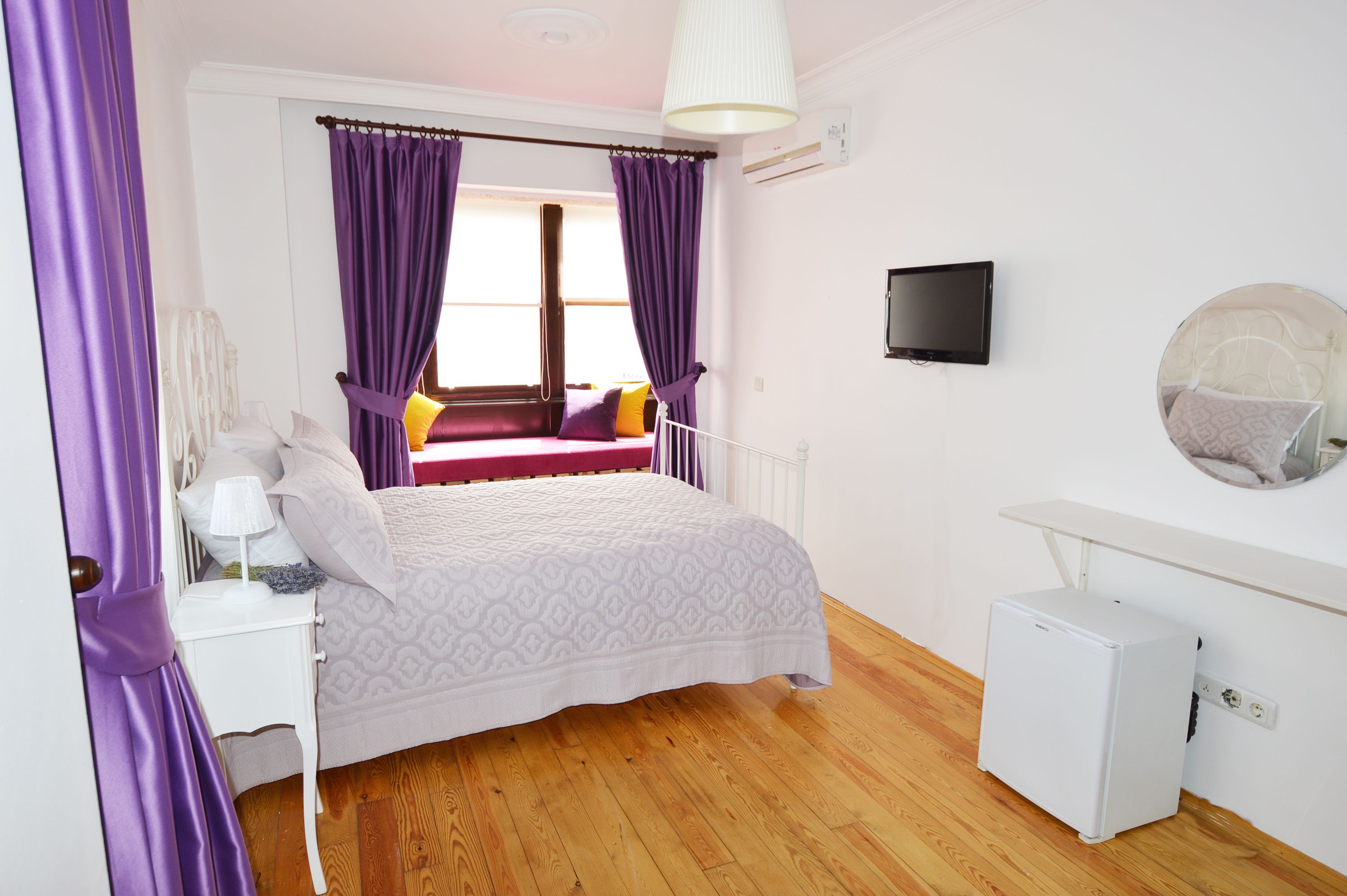 A bedroom with a bed, a small white bedside table, a white nightstand, a wall-mounted TV, a mirror, and a small white heater.