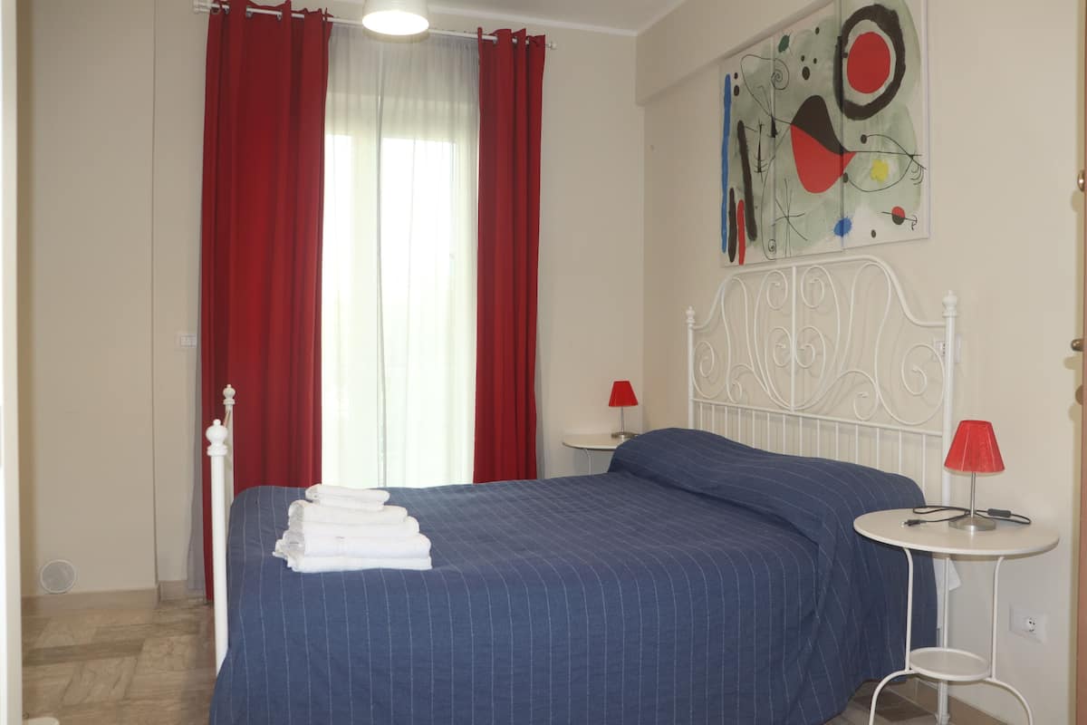Une chambre à coucher avec un lit, des rideaux rouges, une tête de lit blanche, une petite table ronde avec une lampe rouge et une œuvre d’art murale.