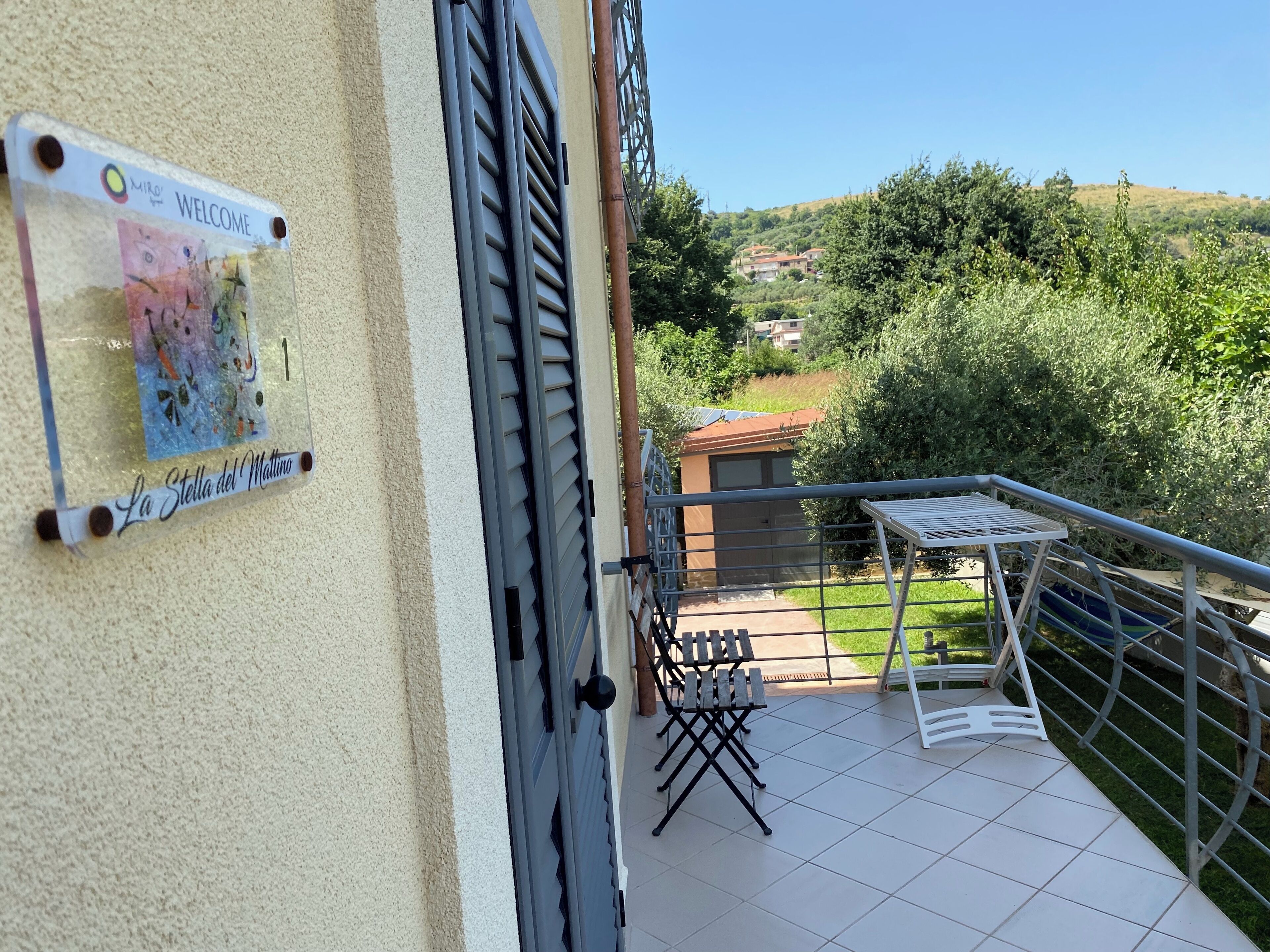 A balcony with a metal railing, a small table, and a chair. There is a sign on the wall with the text "Welcome" and "La Stella del Mattino".