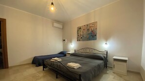 A bedroom with a large bed, two smaller beds, a bedside table, a nightstand, an air conditioning unit, and a wall with abstract art.