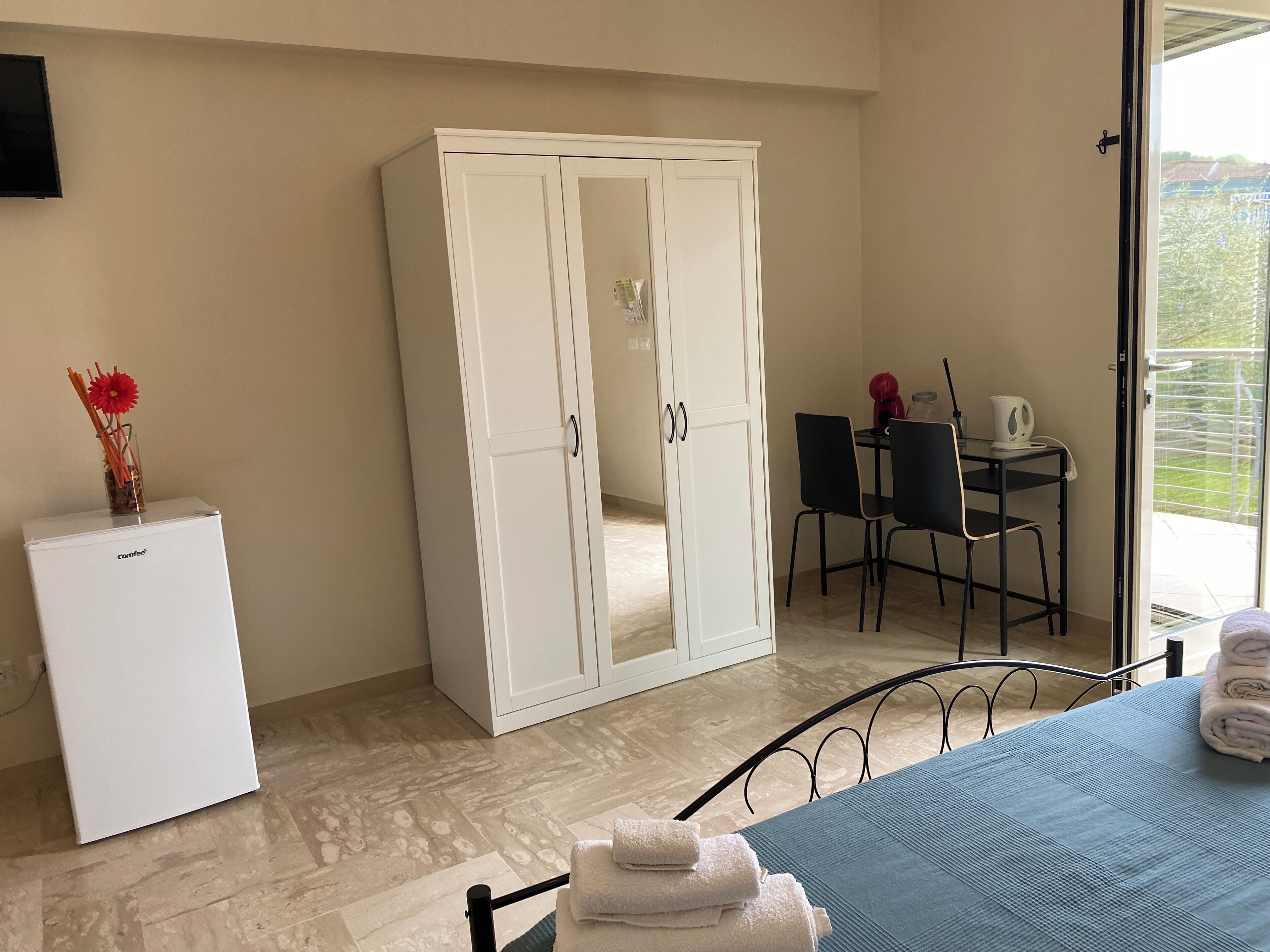 A room with a white wardrobe, a small white fridge, a dining table with chairs, and a view of a balcony.