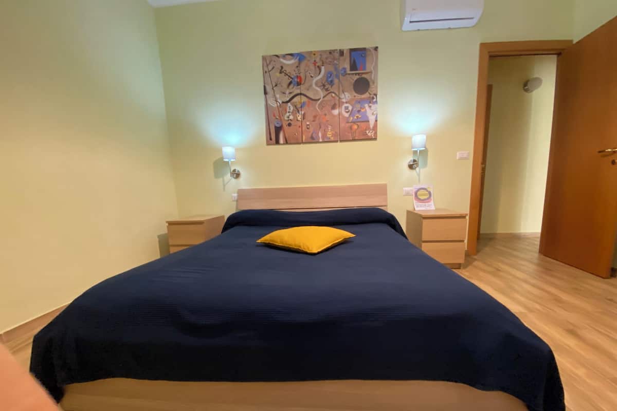Un lit bien fait, recouvert d’une couvre-lit bleue et agrémenté d’un seul oreiller jaune, est flanqué de deux tables de chevet sur lesquelles reposent des lampes. Un tableau abstrait aux couleurs vives orne le mur au-dessus du lit.
