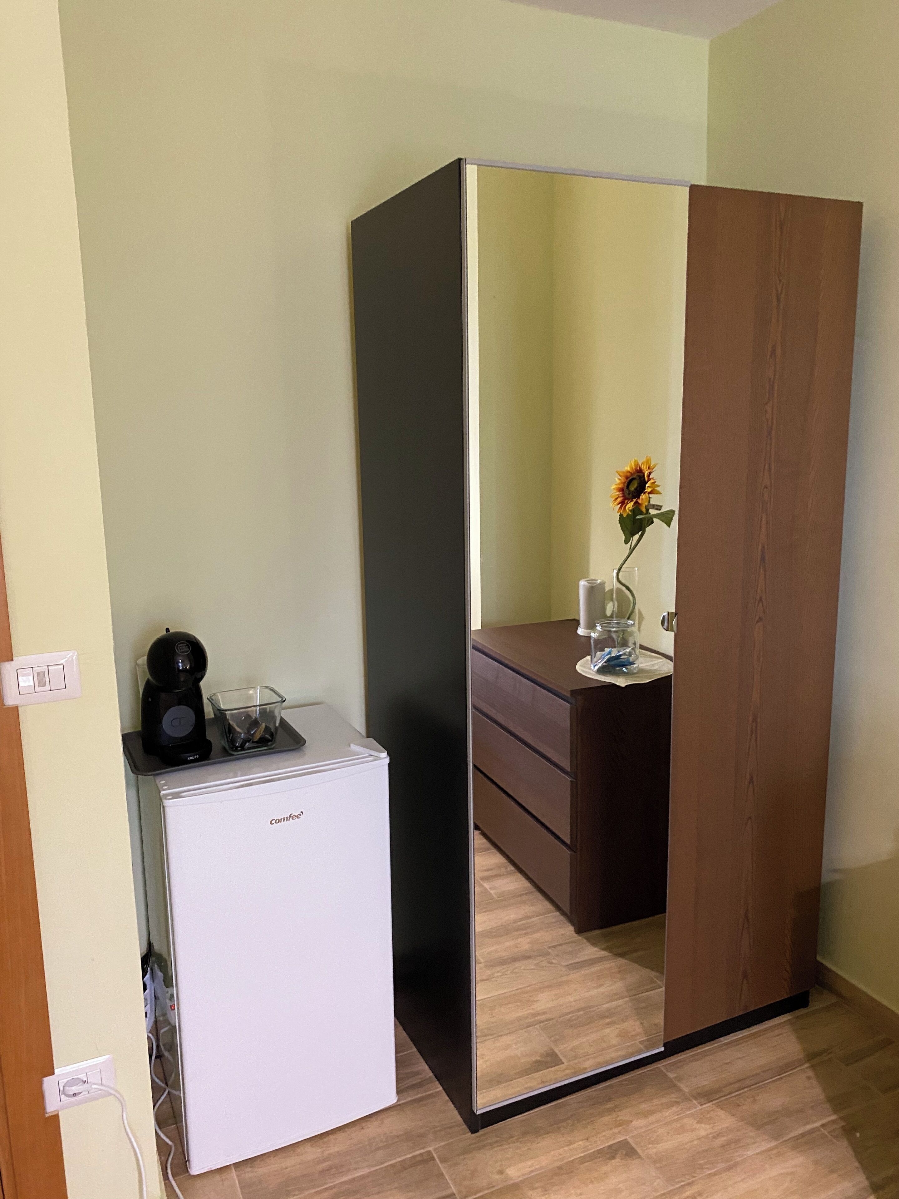 A compact room with a white mini-fridge, a coffee maker, and a small table with a vase of flowers.