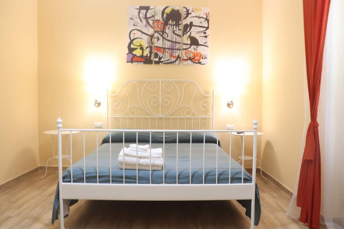 Un lit bien fait, avec un matelas bleu et du linge de lit blanc, une tête de lit en métal blanche et une petite table de chevet avec une lampe.