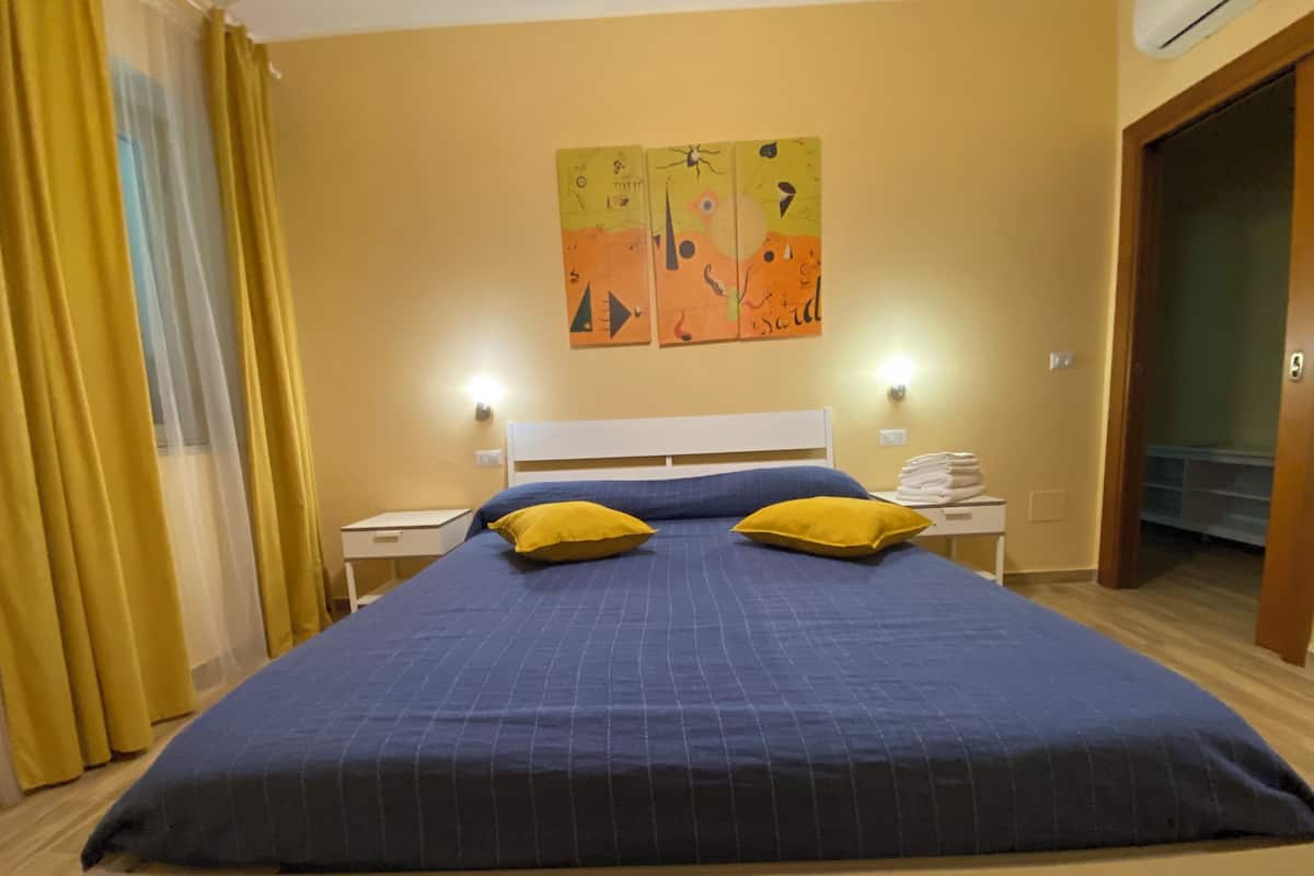 Une chambre à coucher avec un grand lit, deux tables de chevet, une œuvre d’art accrochée au mur et une porte donnant sur une autre pièce.