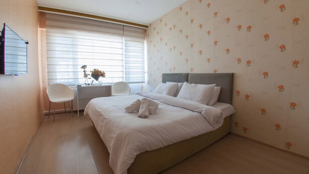 Een keurig opgemaakt bed met witte lakens, een grijs hoofdbord en twee witte kussens. Links staat een klein tafeltje met een bloevas en een stoel. De kamer heeft licht houten vloeren en bloemenbehang.