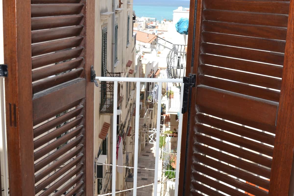 A balcony with wooden shutters, a view of a narrow street, and a distant body of water.