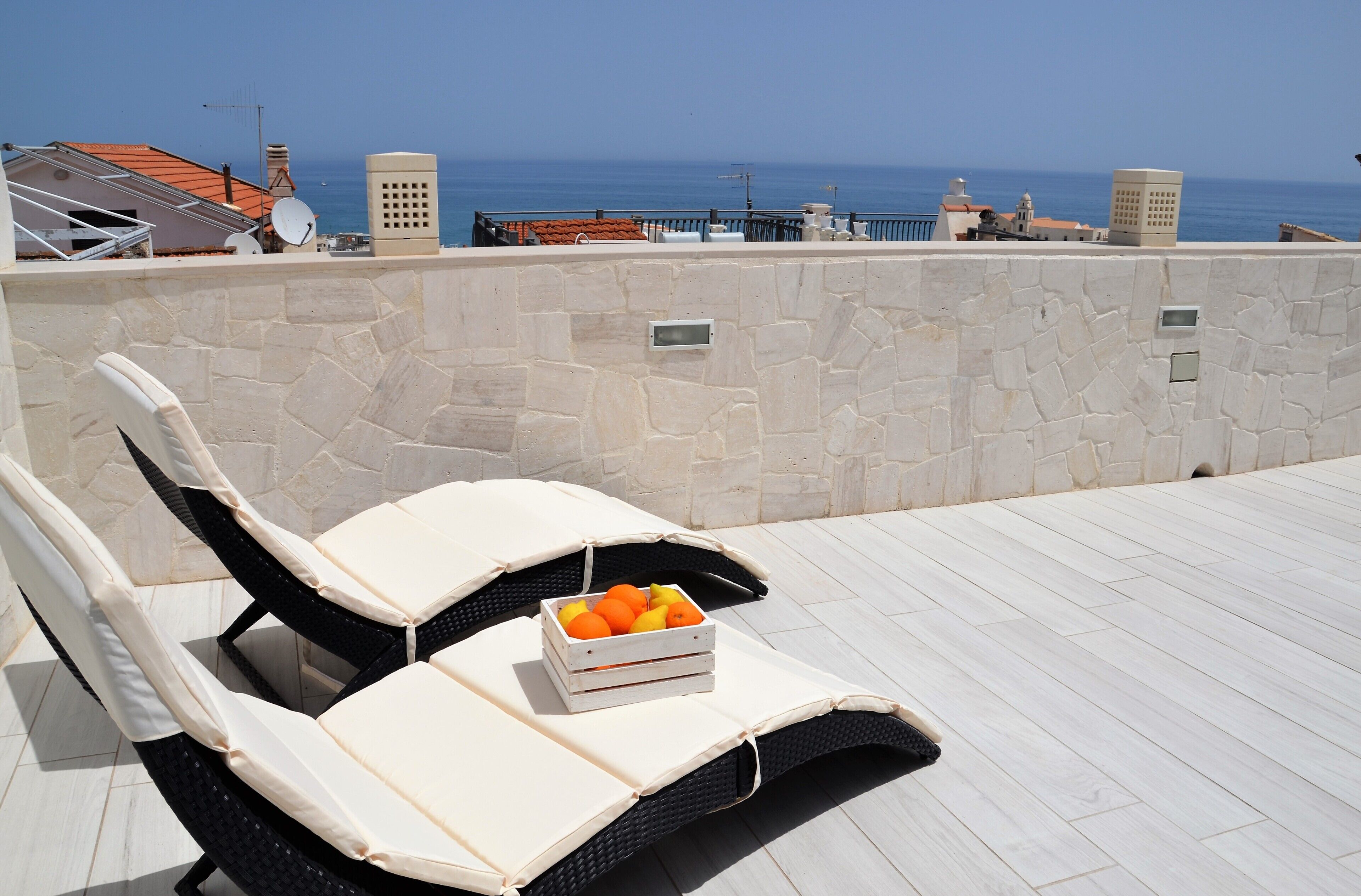 A rooftop lounge with a stone wall, a white cushioned lounge chair, and a crate of oranges.