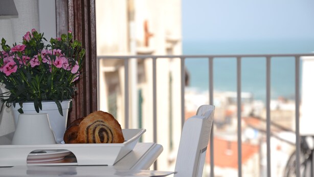 A balcony with a table, a potted plant with pink flowers, and a loaf of bread. - B&B Cuore di Vieste (Vieste)