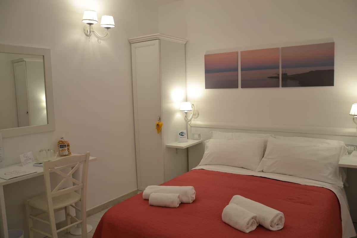 A hotel room with a bed, a desk, a chair, a nightstand, and a wall-mounted lamp.