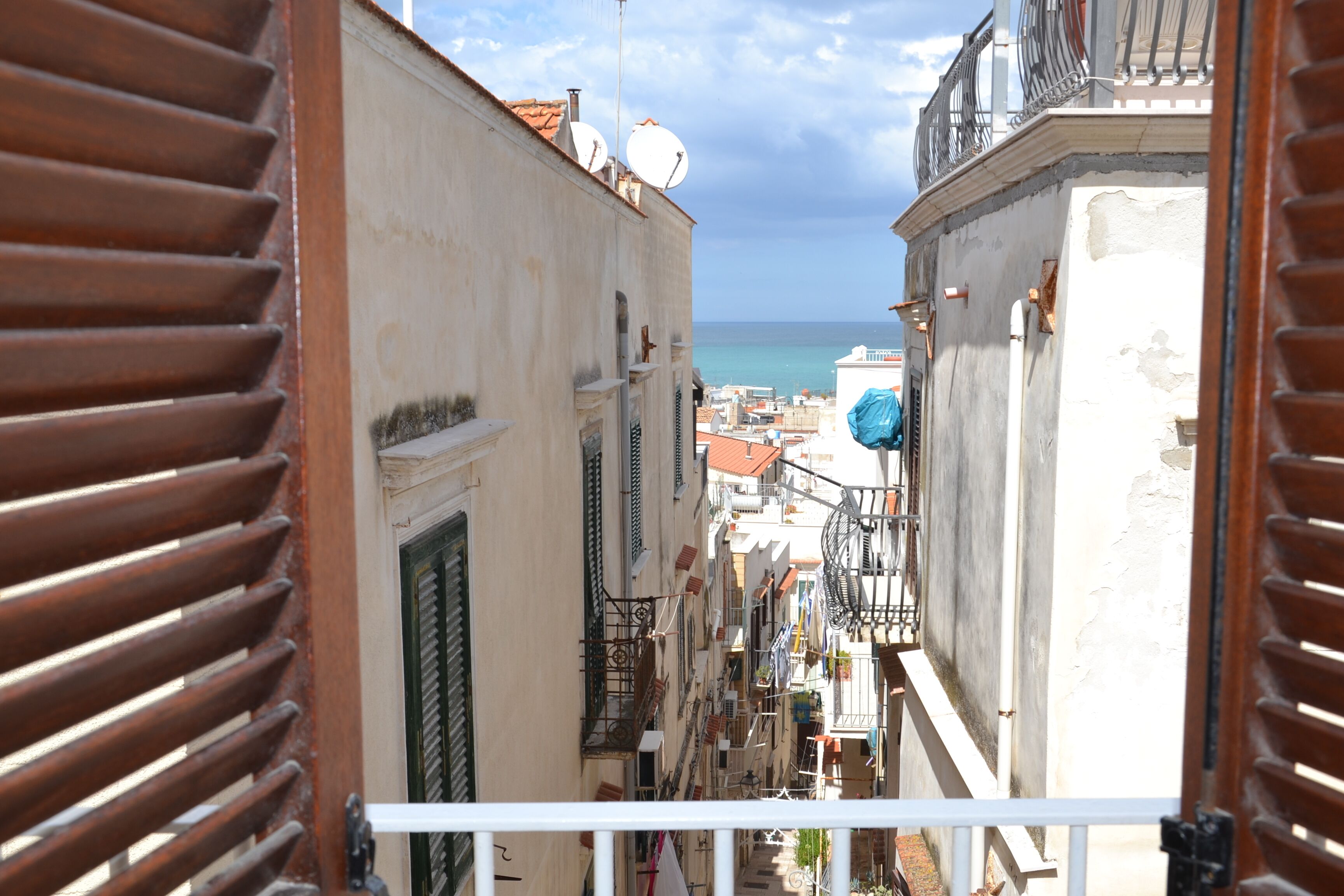 A narrow street with buildings on either side, a view of the sea, and a balcony with a railing.