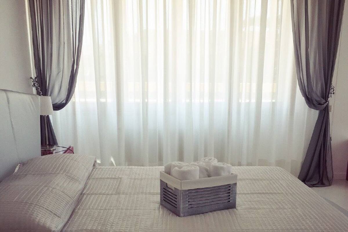A neatly made bed with a gray mattress and pillows, a bedside table with a lamp, and a bedside basket.