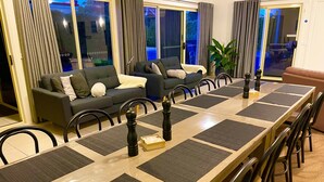 A dining area with a long wooden table, black and grey placemats, and black chairs. There is a grey sofa and a large potted plant in the background. - Austral Place 88 over Merri River (Dennington)