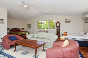 A spacious living room with a ceiling fan, a large window with a view of greenery, a sofa, a coffee table, and a bed with a nightstand.