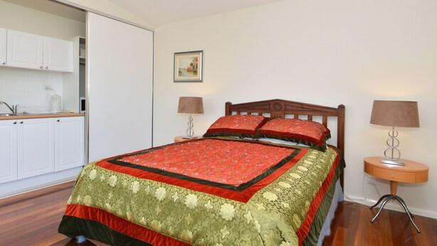 Une chambre à coucher avec un lit, deux lampes de chevet, un tableau accroché au mur et un coin cuisine visible à travers une porte ouverte.