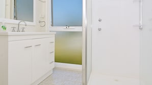Un baño con lavamanos blanco, espejo y mueble, ducha con puerta de cristal y ventana sobre el lavamanos.