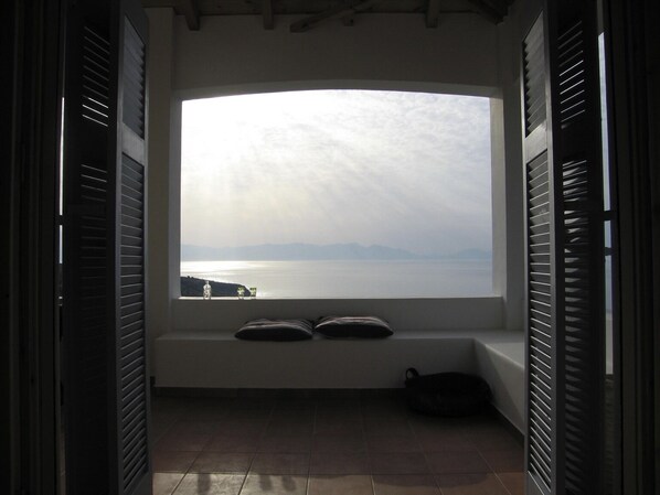 A room with a large window overlooking a body of water and distant mountains. There are two pillows on the windowsill and a bag on the floor.