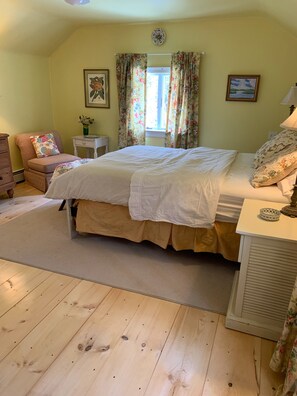 Un dormitorio con cama, una silla, una mesita y una ventana con cortinas florales.