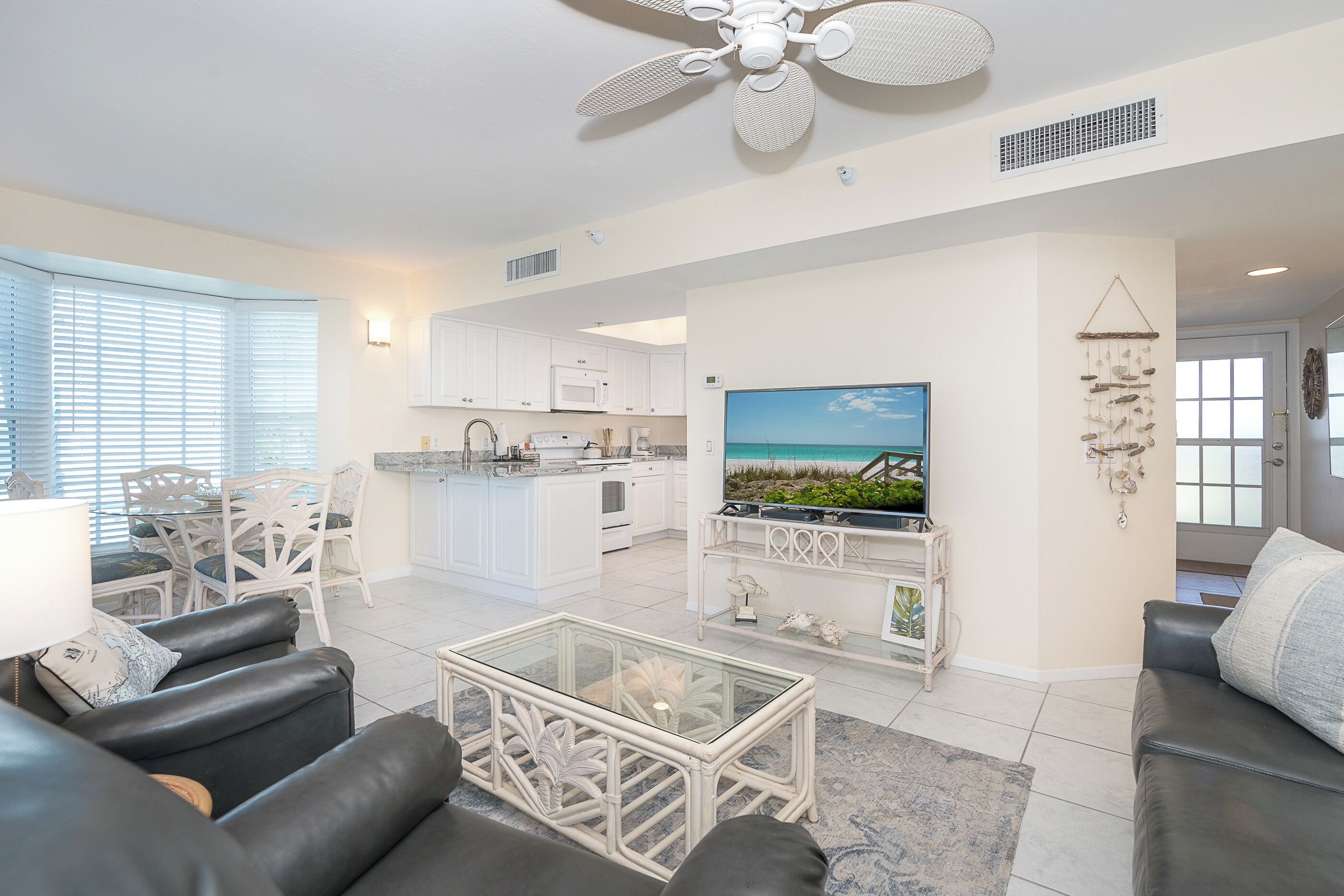 A modern living room with a black leather sofa, a glass coffee table, and a TV on a white stand. There is a kitchen in the background with white cabinets and a dining area with white chairs.