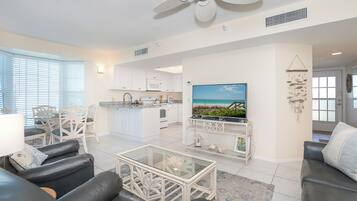 A modern living room with a black leather sofa, a glass coffee table, and a TV on a white stand. There is a kitchen in the background with white cabinets and a dining area with white chairs.