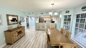A spacious dining area with a wooden table and chairs, a kitchen in the background, and a view through large windows. - Perfect for a Beach Getaway, Great View and Near the Pool, B2613A (Cape Haze)