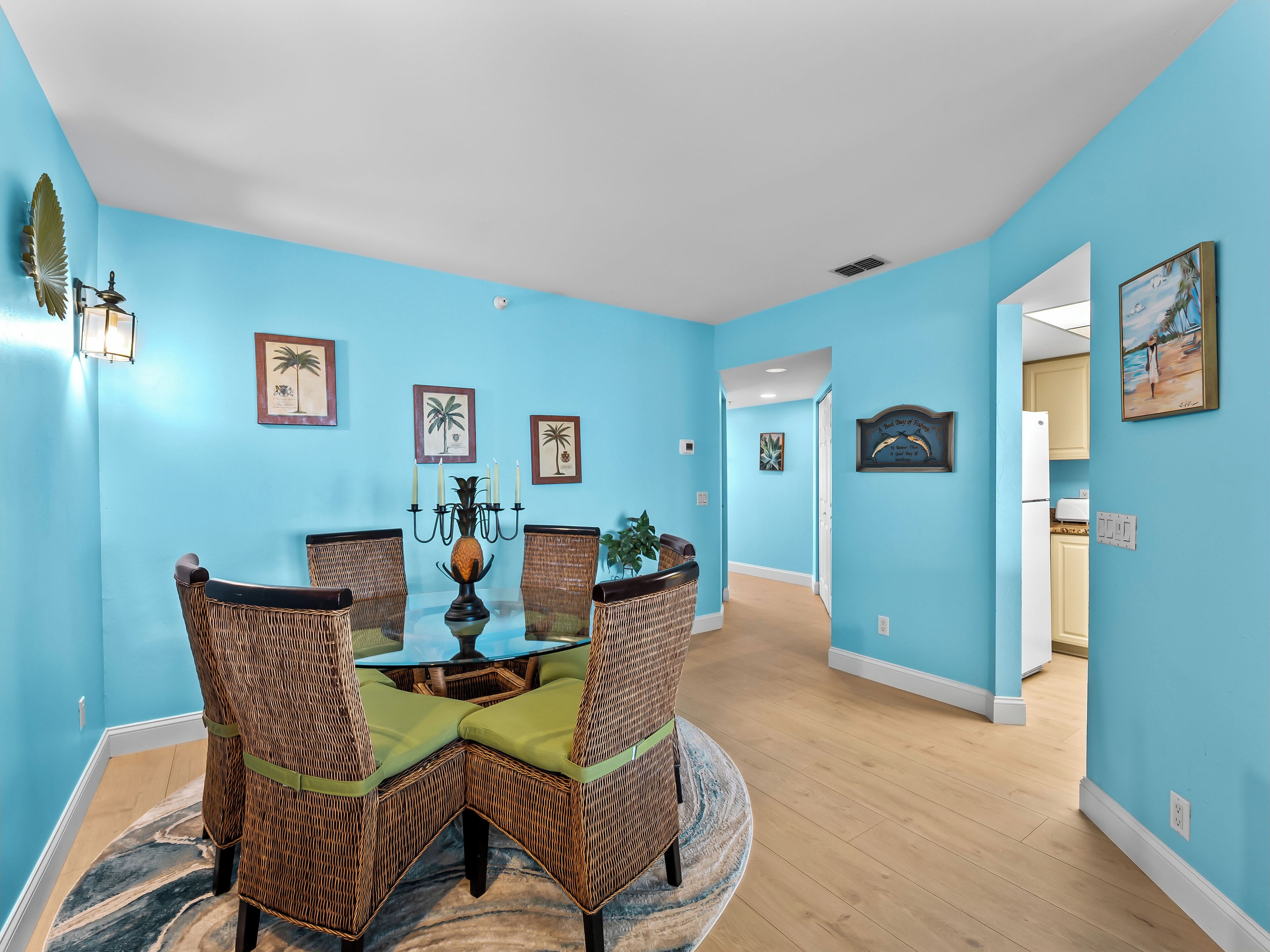 A dining area with a round table and four wicker chairs, a chandelier, and framed artwork on the walls.
