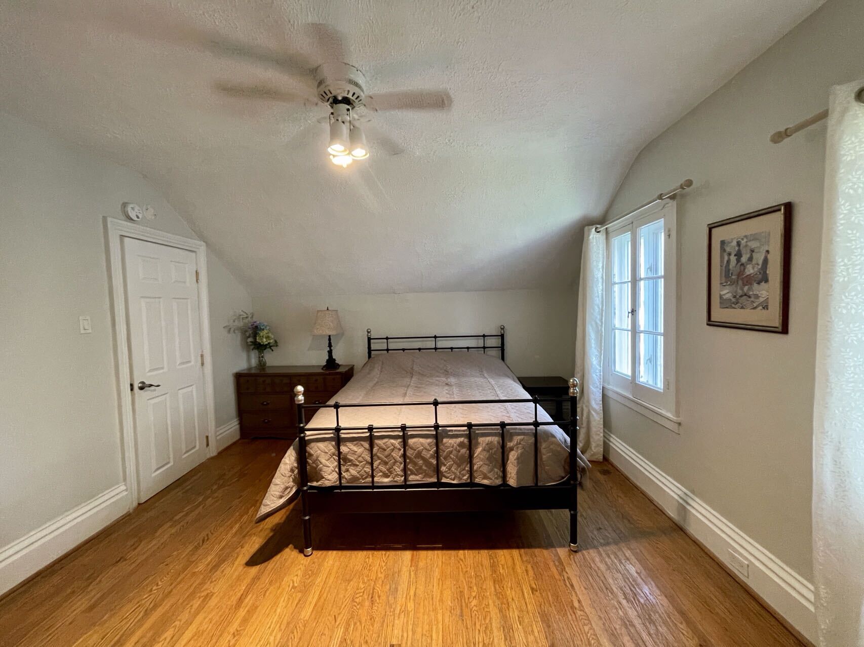 A bedroom with a bed, a ceiling fan, a nightstand, a lamp, a window, and a door.