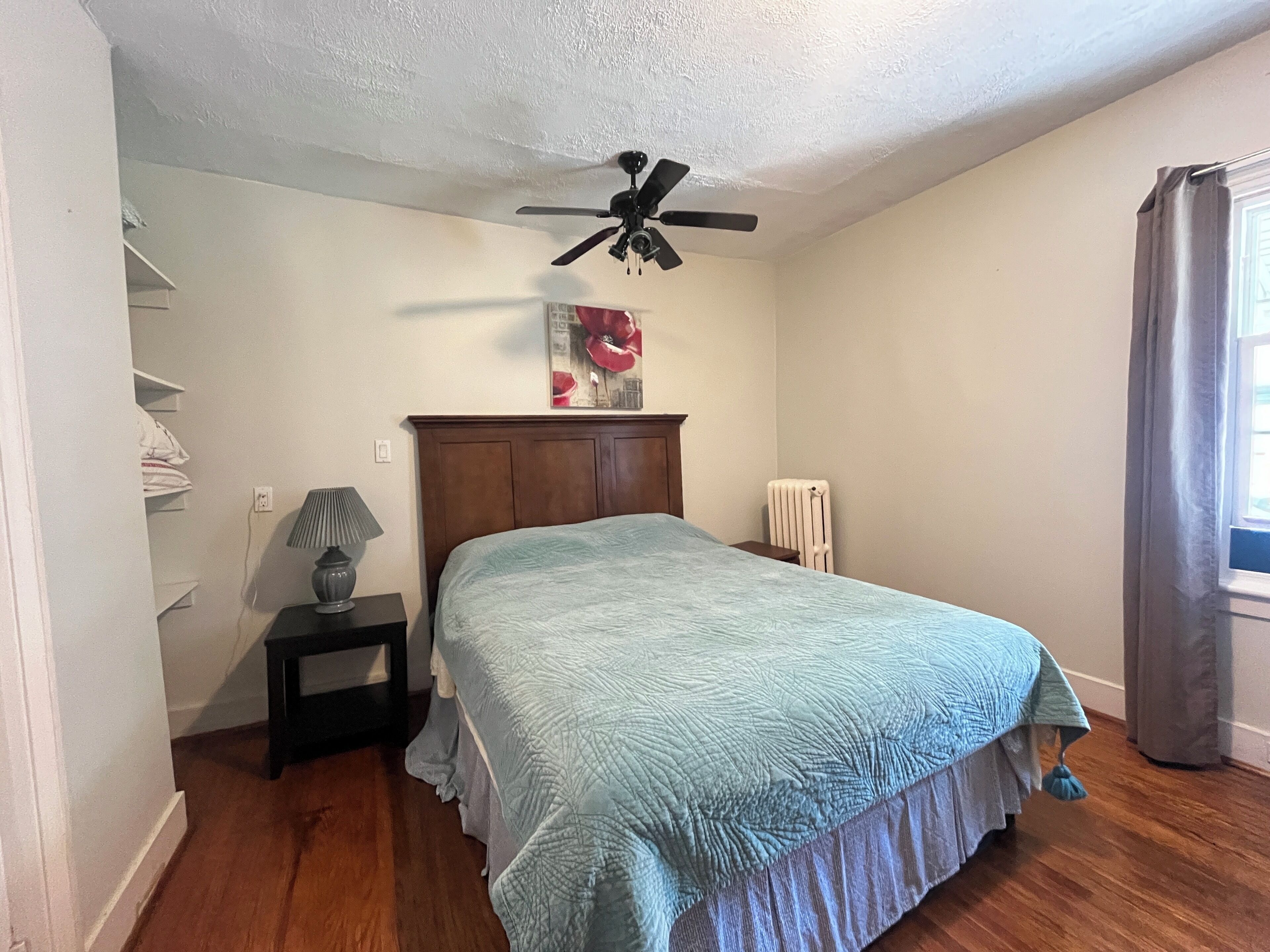 A bedroom with a bed, a ceiling fan, a radiator, and a window with curtains.