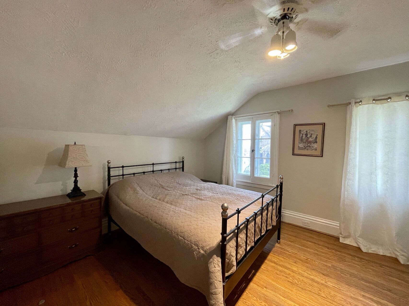 A bedroom with a bed, a dresser, a window, a ceiling fan, and a painting on the wall.