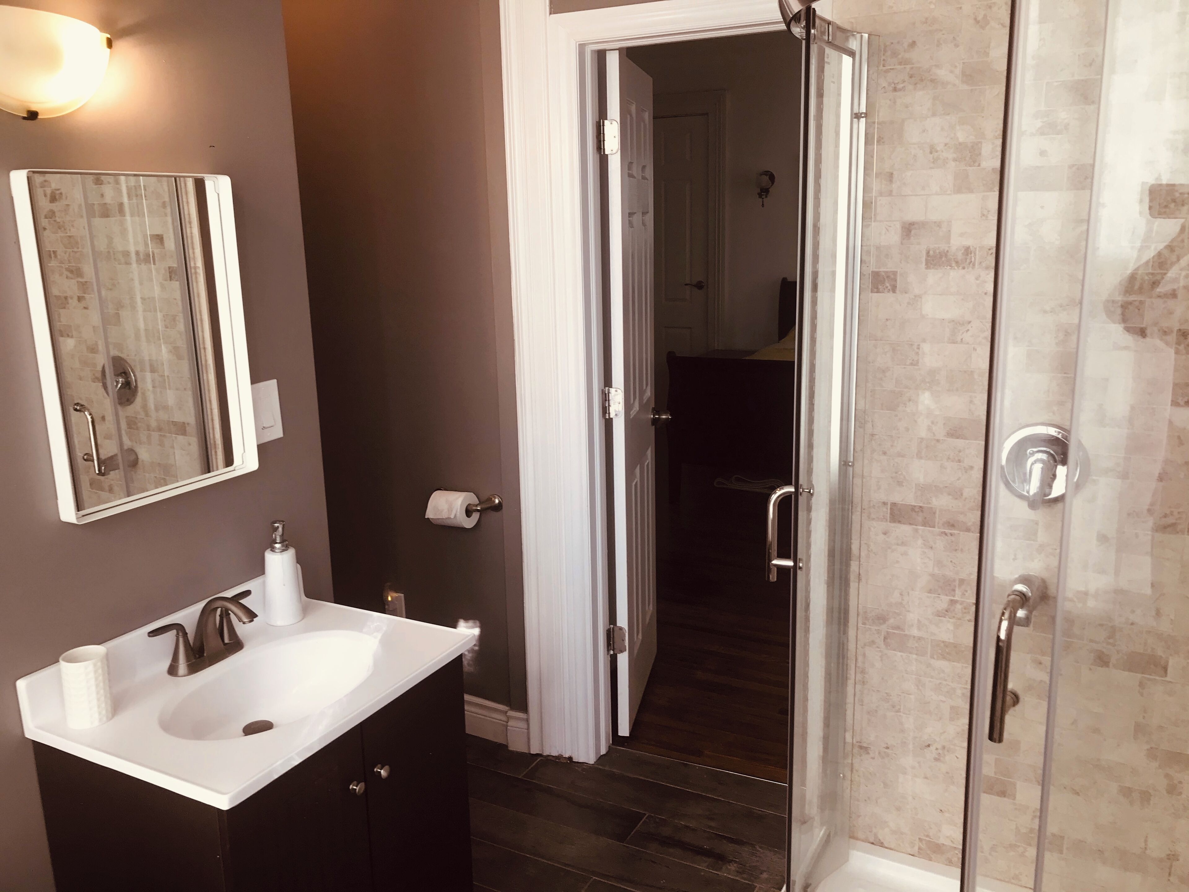 Un baño con un lavamanos blanco, muebles oscuros, una cabina de ducha de vidrio y una puerta que da a otra habitación.
